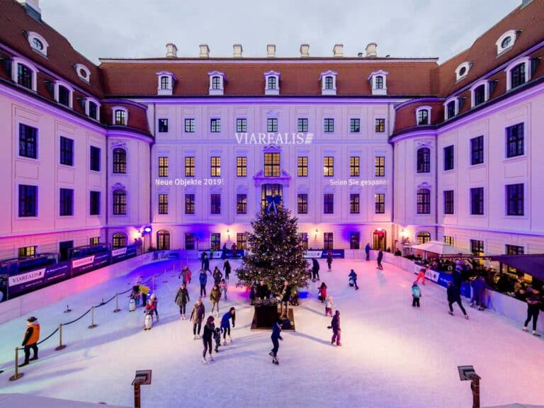 Ein belebter Innenhof mit Menschen, die um einen großen, geschmückten Weihnachtsbaum Schlittschuh laufen. Das umliegende, lila beleuchtete Gebäude ist mit Schildern versehen, die auf „Immobilienmakler Dresden“ hinweisen. Festliche Lichter schmücken den Bereich und sorgen für eine lebendige Winteratmosphäre.