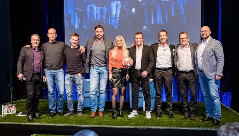 Eine Gruppe von neun Personen steht auf einer grasähnlichen Bühne, einer hält einen Fußball in der Hand. In legerer Business-Kleidung verkörpern sie Professionalität. Hinter ihnen zeigt ein großer Bildschirm ein digitales Bild, möglicherweise aus einer Immobilienberatung oder Tipps für die Beratung beim Immobilienkauf.