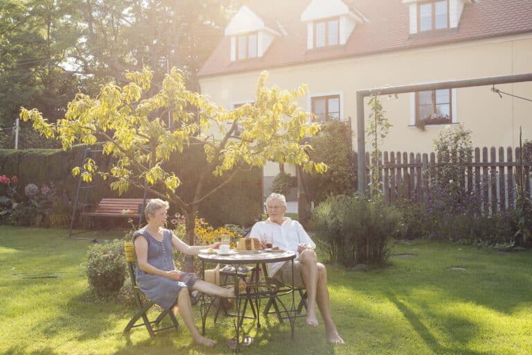 Ein älteres Paar sitzt an einem runden Tisch in einem sonnigen Garten, im Schatten eines kleinen Baumes. Lächelnd und bei einem Drink besprechen sie die Aussicht auf einen Hauskauf in Dresden. Die friedliche Umgebung mit einem Haus und einer Gartenschaukel im Hintergrund spiegelt ihre Vorfreude auf den Neuanfang wider.