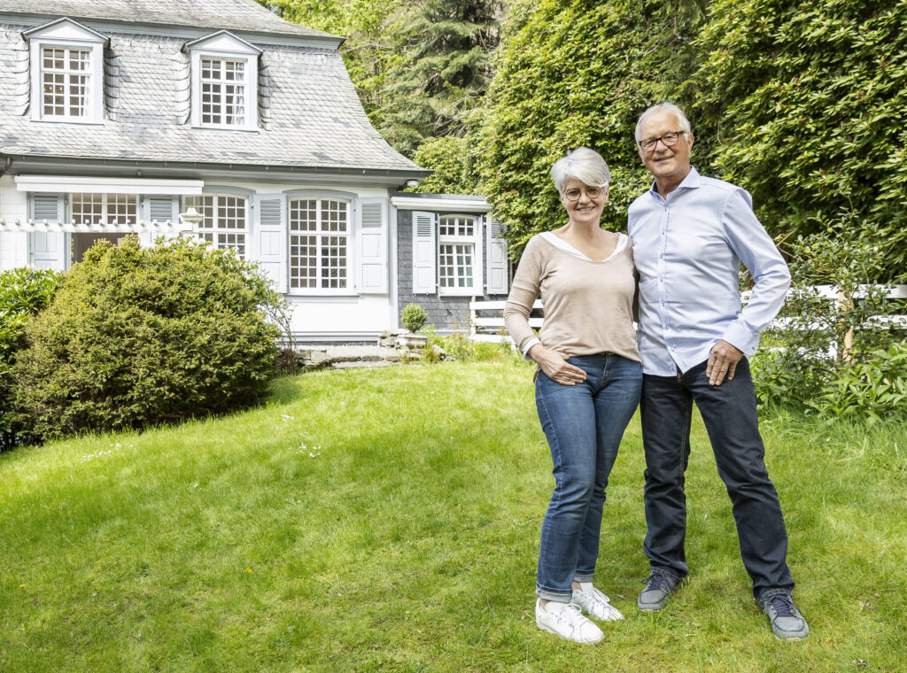 Ein lächelndes älteres Paar steht vor einem charmanten weißen Haus mit grauem Dach, umgeben von üppigem Grün. Die Frau trägt ein beiges Top und Jeans, der Mann ein hellblaues Hemd und Jeans. Sie stehen auf einem gepflegten Rasen.