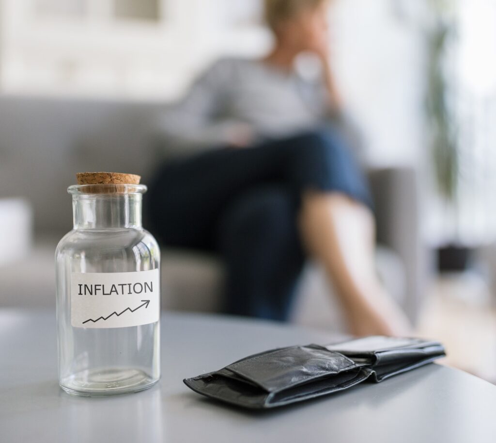 Ein Glas mit der Aufschrift „INFLATION“ und einem nach oben gerichteten Pfeil steht auf einem Tisch neben einer leeren Brieftasche. Im Hintergrund wirkt eine Person nachdenklich.