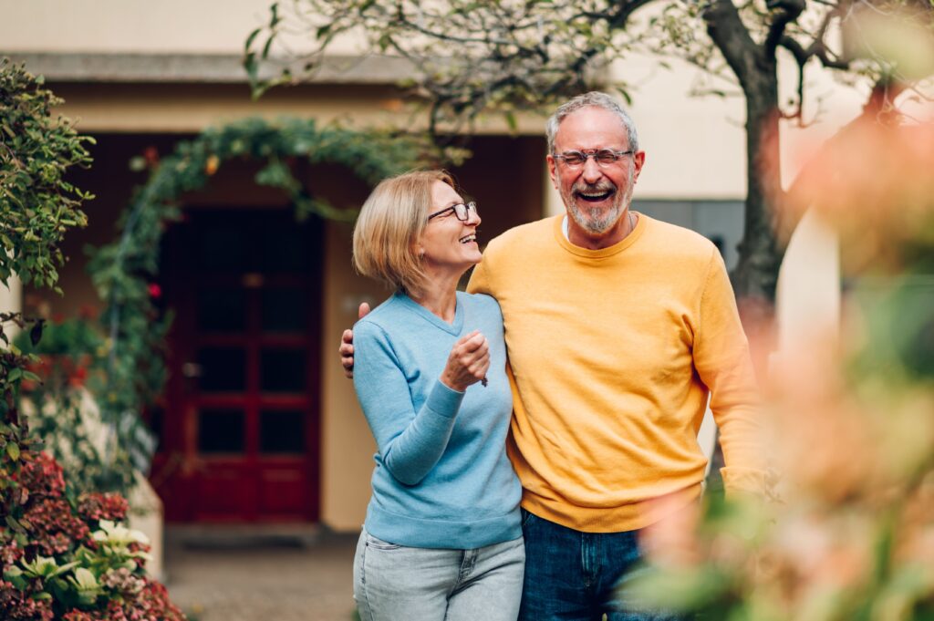 Ein älteres Paar steht Arm in Arm im Garten ihres Hauses. In entspannter Atmosphäre denken sie darüber nach, ob das Wohnen in der eigenen Immobilie oder eine vermietete Wohnung die bessere Lösung für den Ruhestand ist. Was bringt mehr – das Eigenheim oder die Kapitalanlage zur Altersvorsorge?
