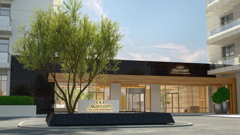 Moderne Außenansicht des Eingangs der Marriott Executive Apartments mit großen Glastüren, einer schwarzen Steinmauer mit dem Marriott-Logo, einem begrünten Pflanzgefäß mit einem Baum und nahe gelegenen Gebäuden.