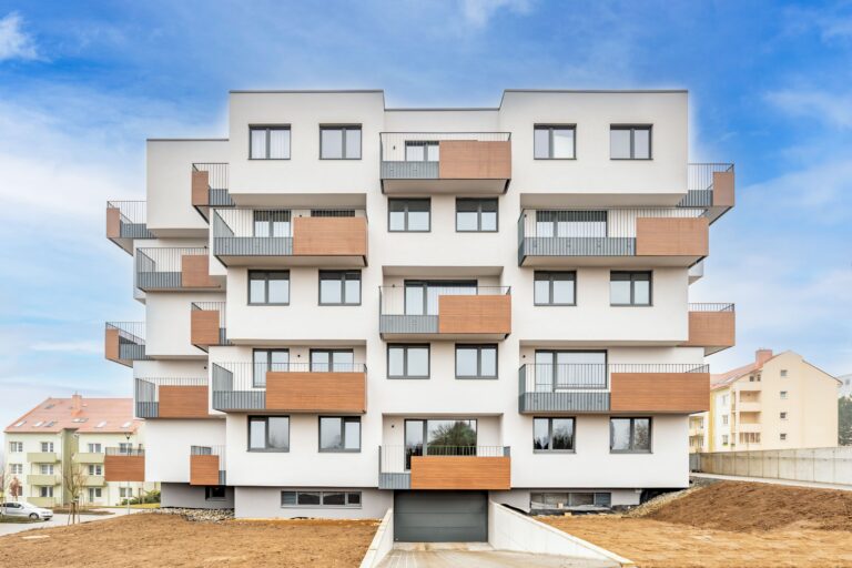 Modern white apartment building with several balconies with wooden cladding and metal railings against a blue sky. An unfinished floor area and a ramp to the garage can be seen in the foreground - ideal for anyone looking to buy a home.