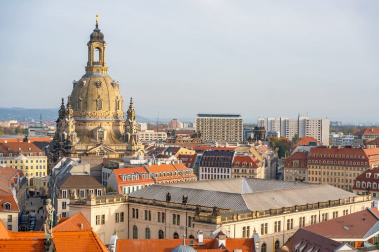 Ein Panoramablick auf Dresden, Deutschland, mit der Kuppel der Frauenkirche, umgeben von historischen Gebäuden mit roten Dächern und modernen Bauten im Hintergrund.