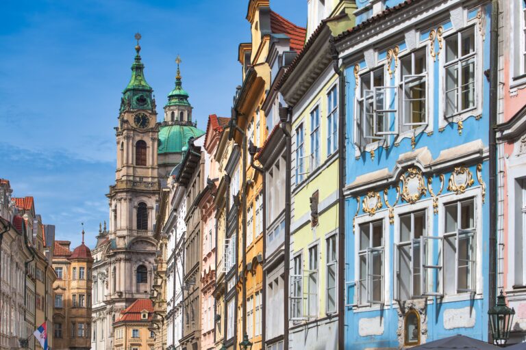 Farbenfrohe historische Gebäude mit kunstvollen Details säumen eine Straße; im Hintergrund ragt ein Kirchturm mit grüner Kuppel vor dem blauen Himmel empor.