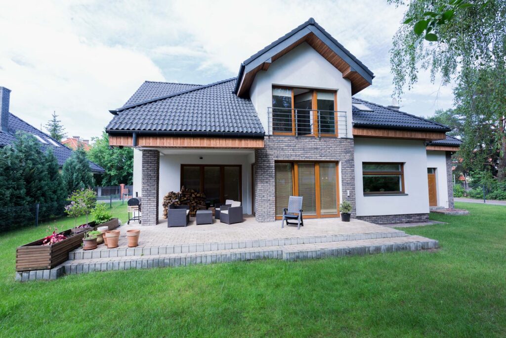 Modern two-story house with a dark roof, light walls, and brick accents. There is a patio with potted plants, chairs, and stacked firewood, surrounded by green lawn and trees.
