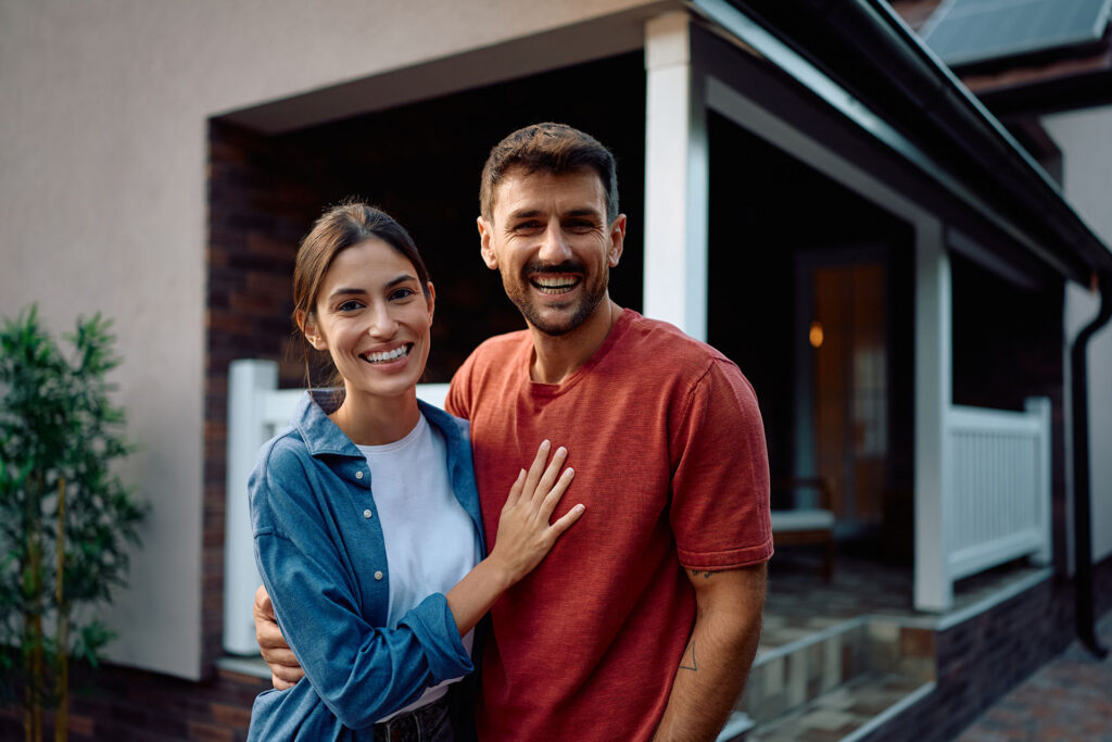 A smiling couple stands in front of a house. The woman places her hand on the man's chest while he has his arm around her. Both look happy, and the house's porch is visible behind them.