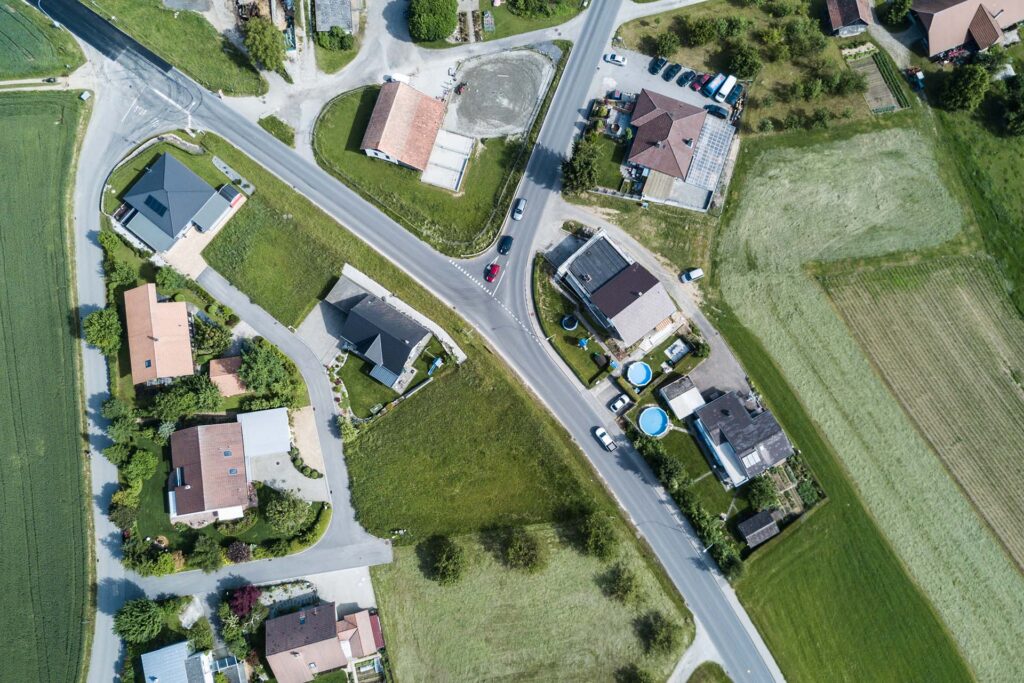 Aerial view of a small town intersection with houses, green lawns, parked cars, gardens, and round above-ground pools, surrounded by fields and trees on a sunny day.