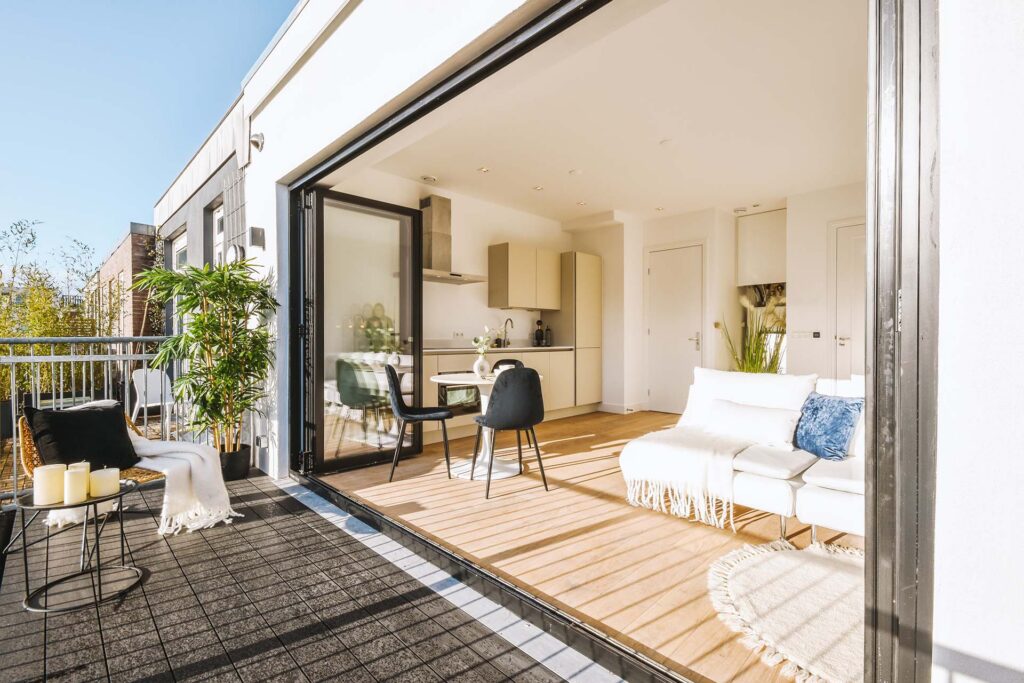 Modern apartment with large, open sliding doors leading to a sunny balcony. Indoors, there is a small dining table, a white sofa, a kitchen, and decorative items. Outdoors, there are chairs, a small table, candles, and plants.