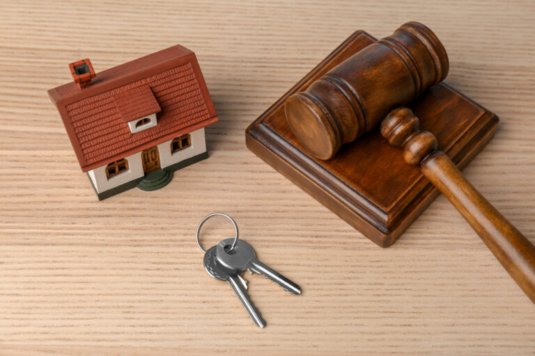 Ein Miniaturhausmodell, ein Holzhammer auf einem Klangblock und ein Schlüsselbund auf einer hellen Holzfläche, die die Spekulationsfrist von „Immobilien 2026“ symbolisieren.